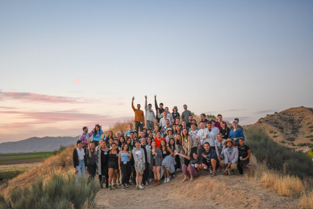 1st night photo 3 A large group of graduate students poses together on a dirt hill at sunset, surrounded by desert plants and distant mountains under a colorful sky.