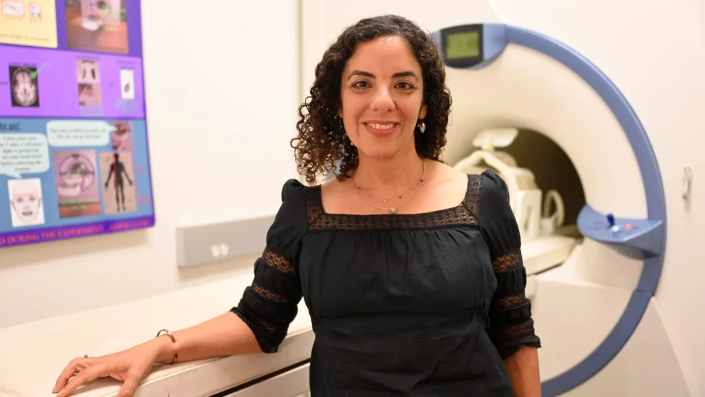 Lisa Aziz-Zadeh stands smiling in front of an fMRI scanner, wearing a black blouse with lace details.