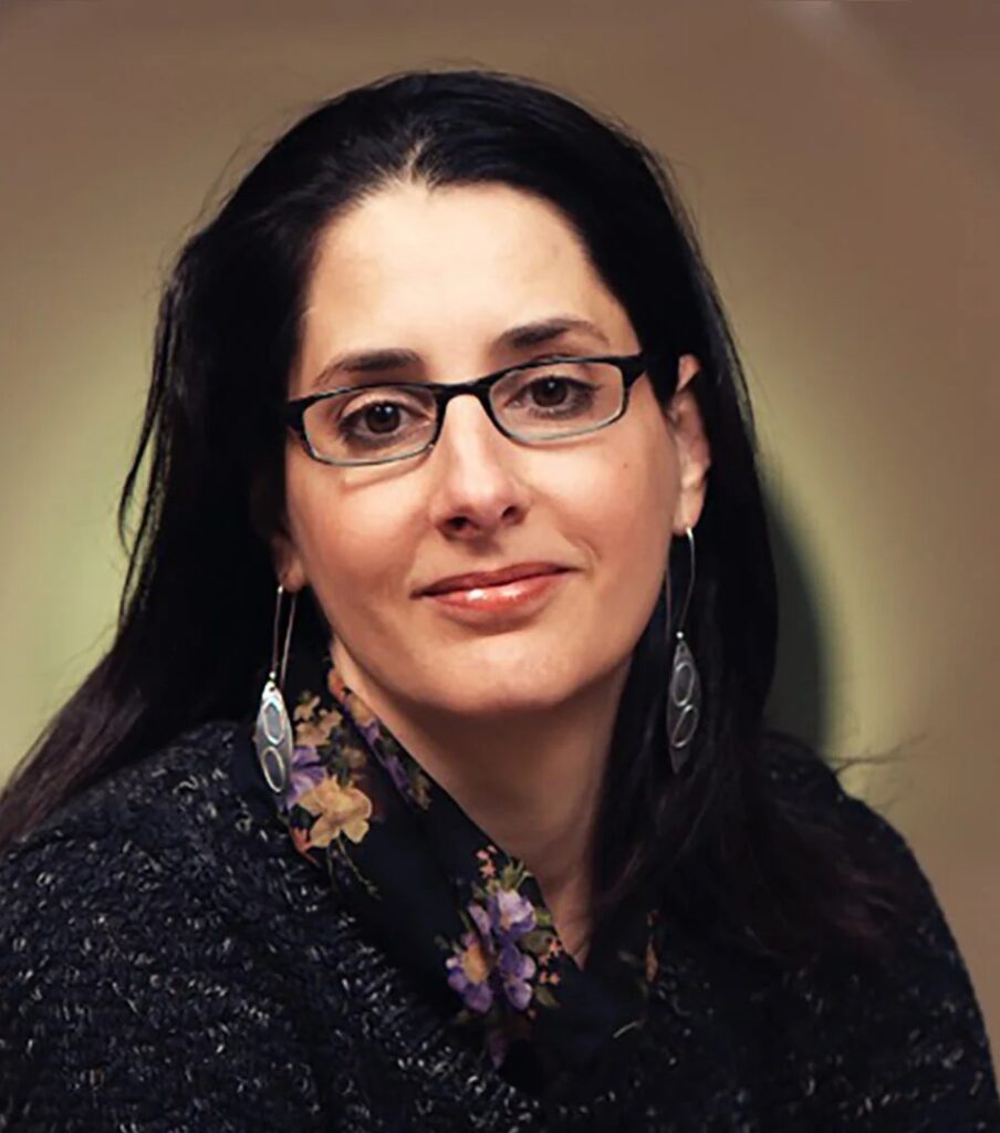 Mary Helen Immordino-Yang, wearing glasses, long earrings, and a floral scarf, poses against a softly lit background.
